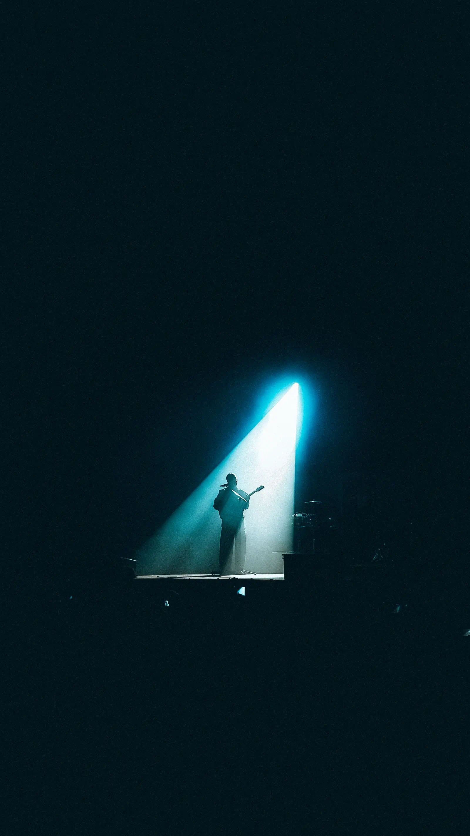 A guitarist alone on a dark stage inside a single cone of teal light