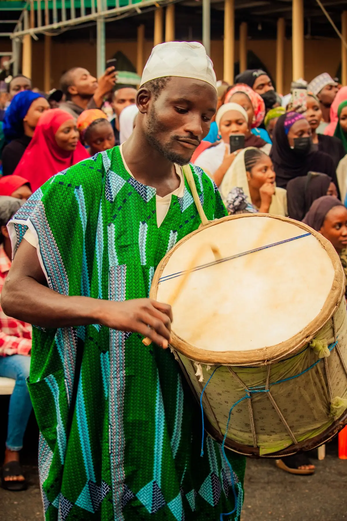 A drummer in a green robe playing a large hand drum in a crowd