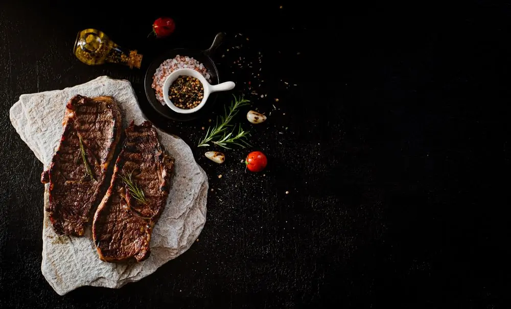 A raw seasoned steak laid on dark slate with rosemary and pepper