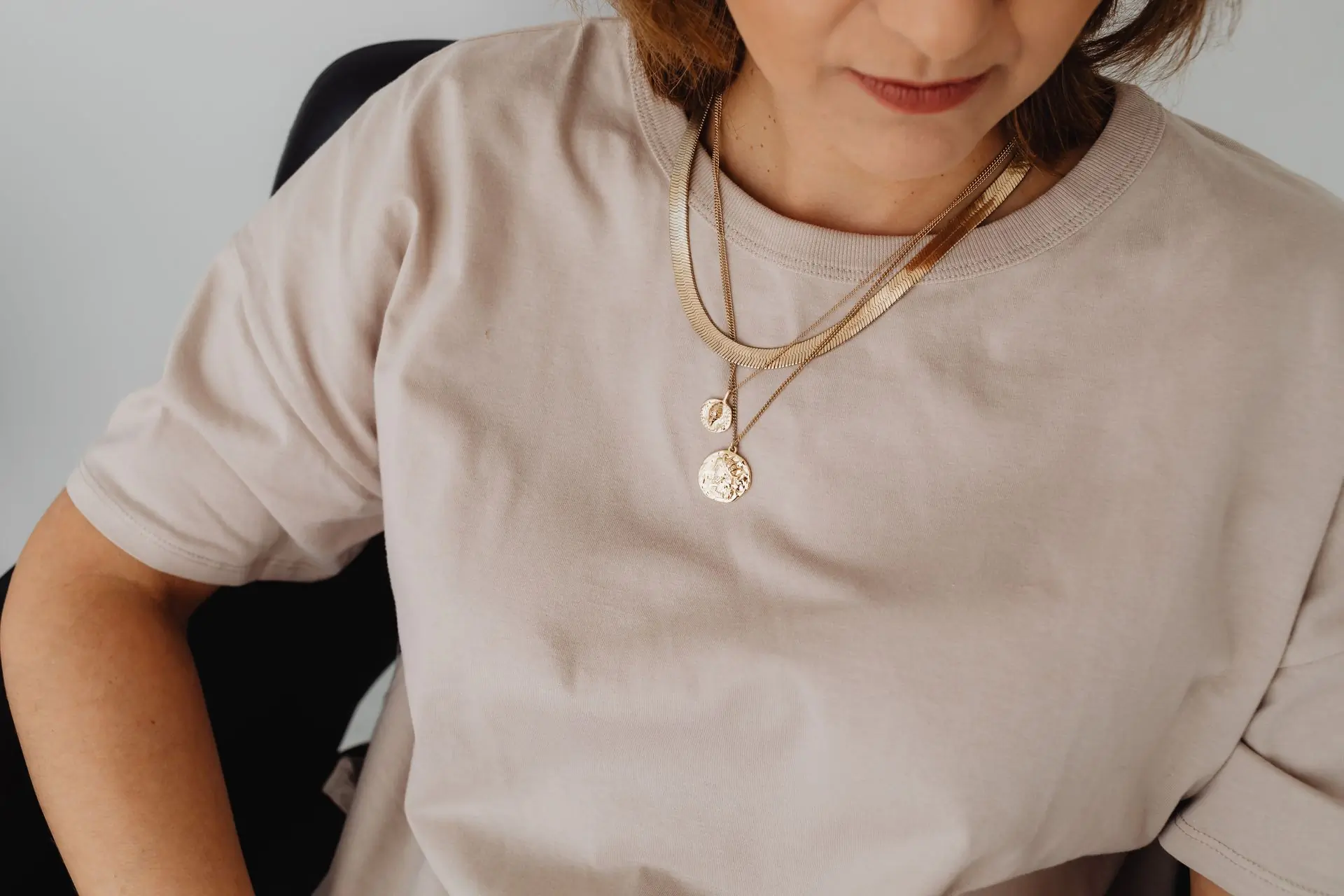 A woman in a blush tee wearing layered gold chain and pendant necklaces against a white studio wall