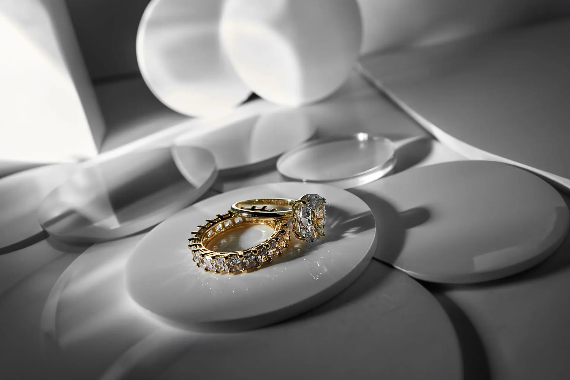 A gold diamond eternity band and solitaire ring staged on pale sculpted discs in soft daylight