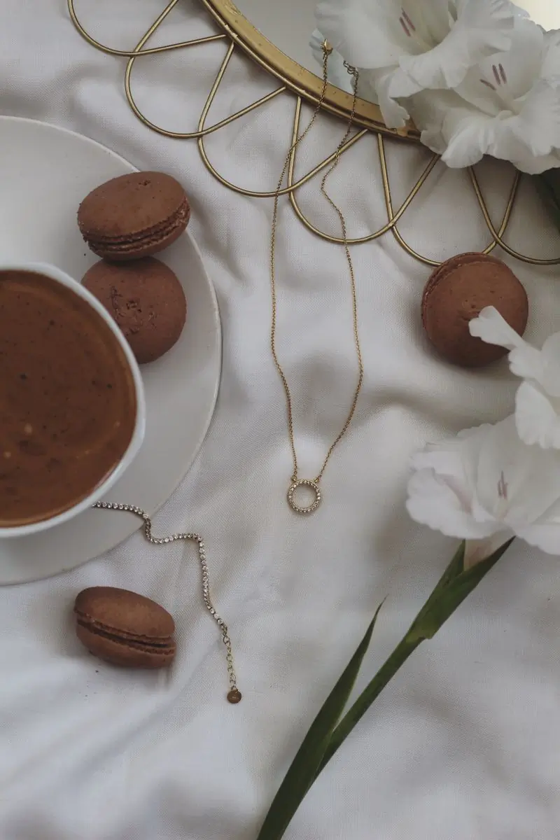 Coffee, macarons and white flowers laid out with a gold chain necklace on cream linen