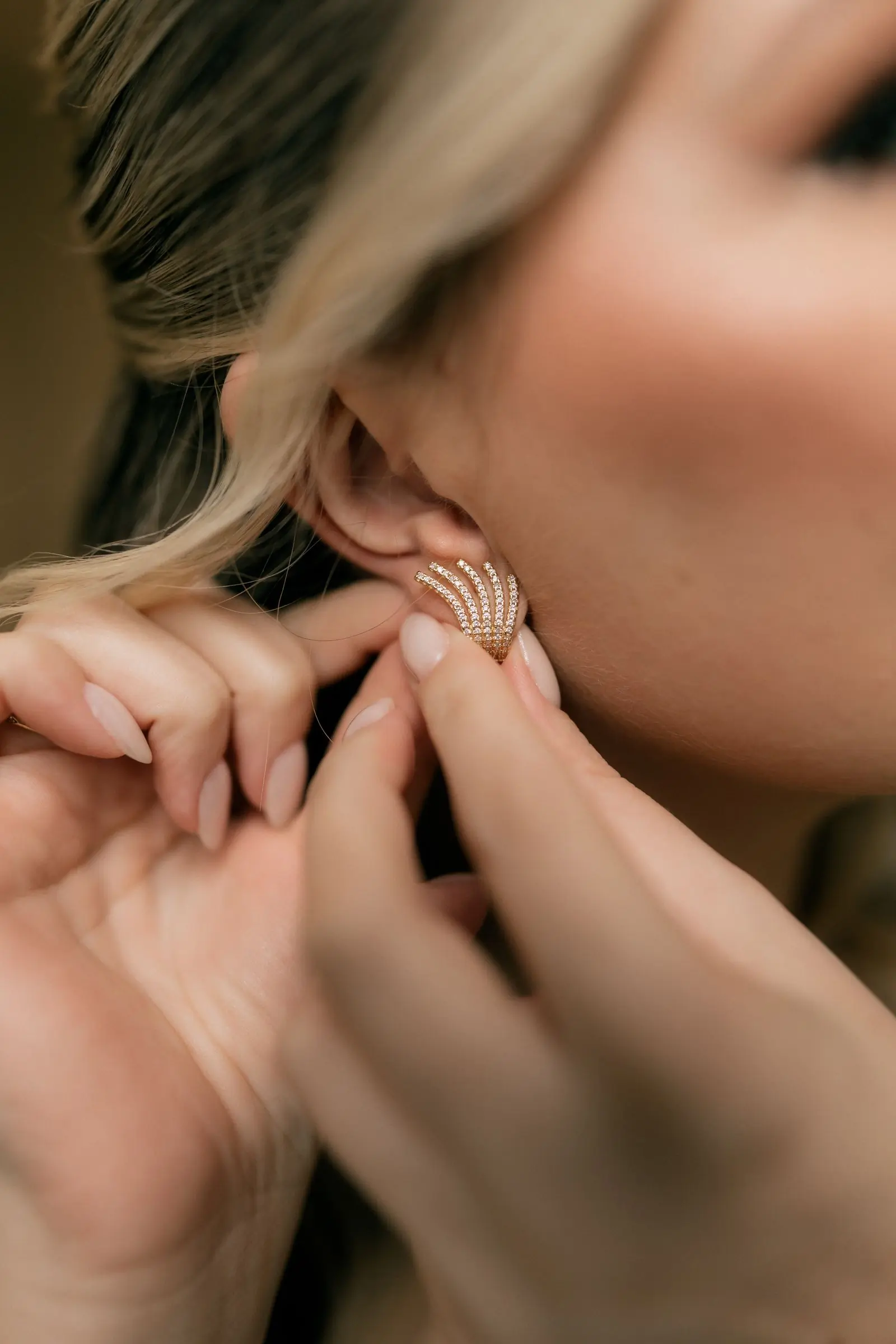 A woman's hands fastening a delicate gold earring at her ear in warm beige light