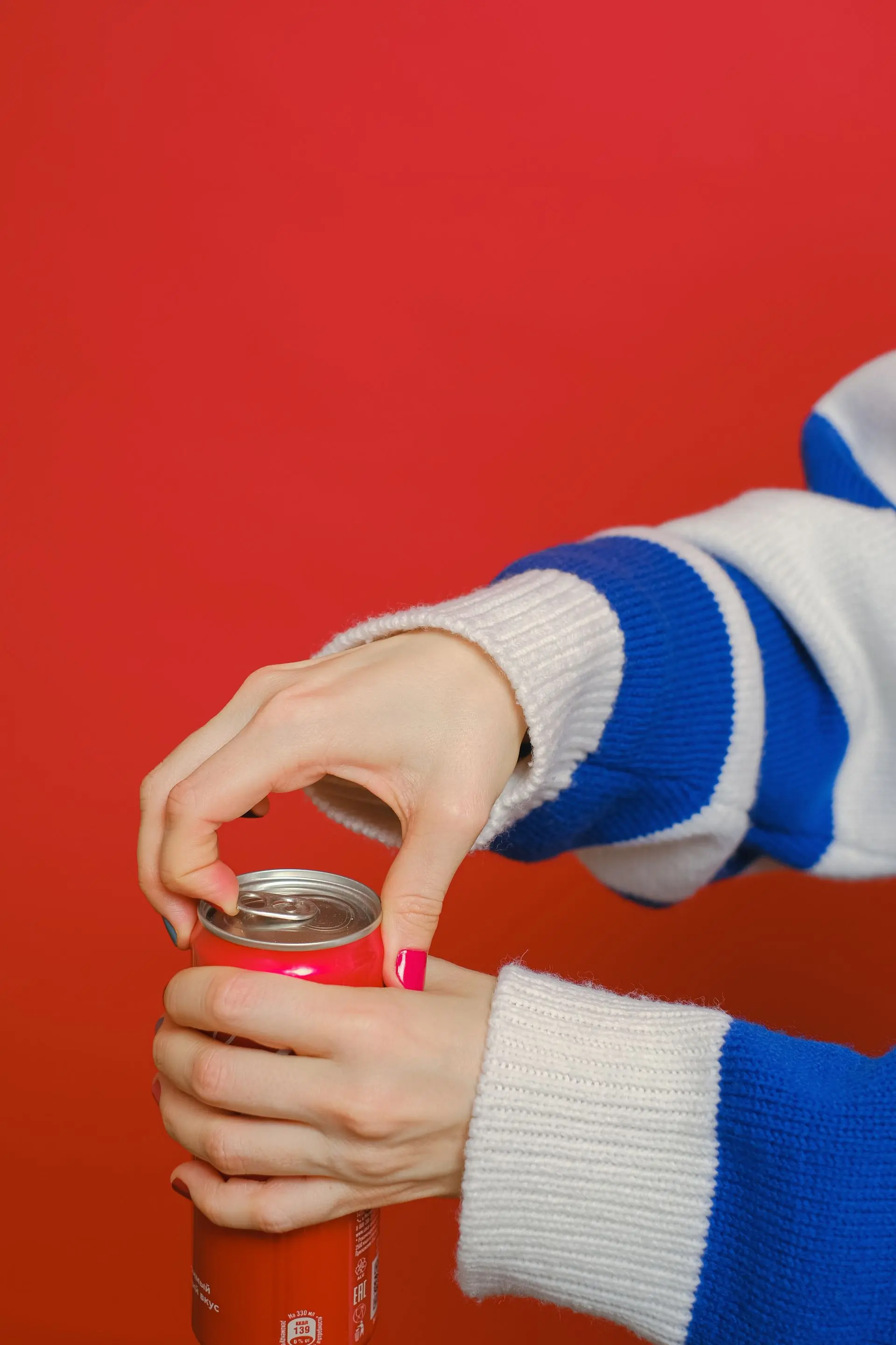 A hand in a striped sleeve opening a red can against a saturated red studio ground