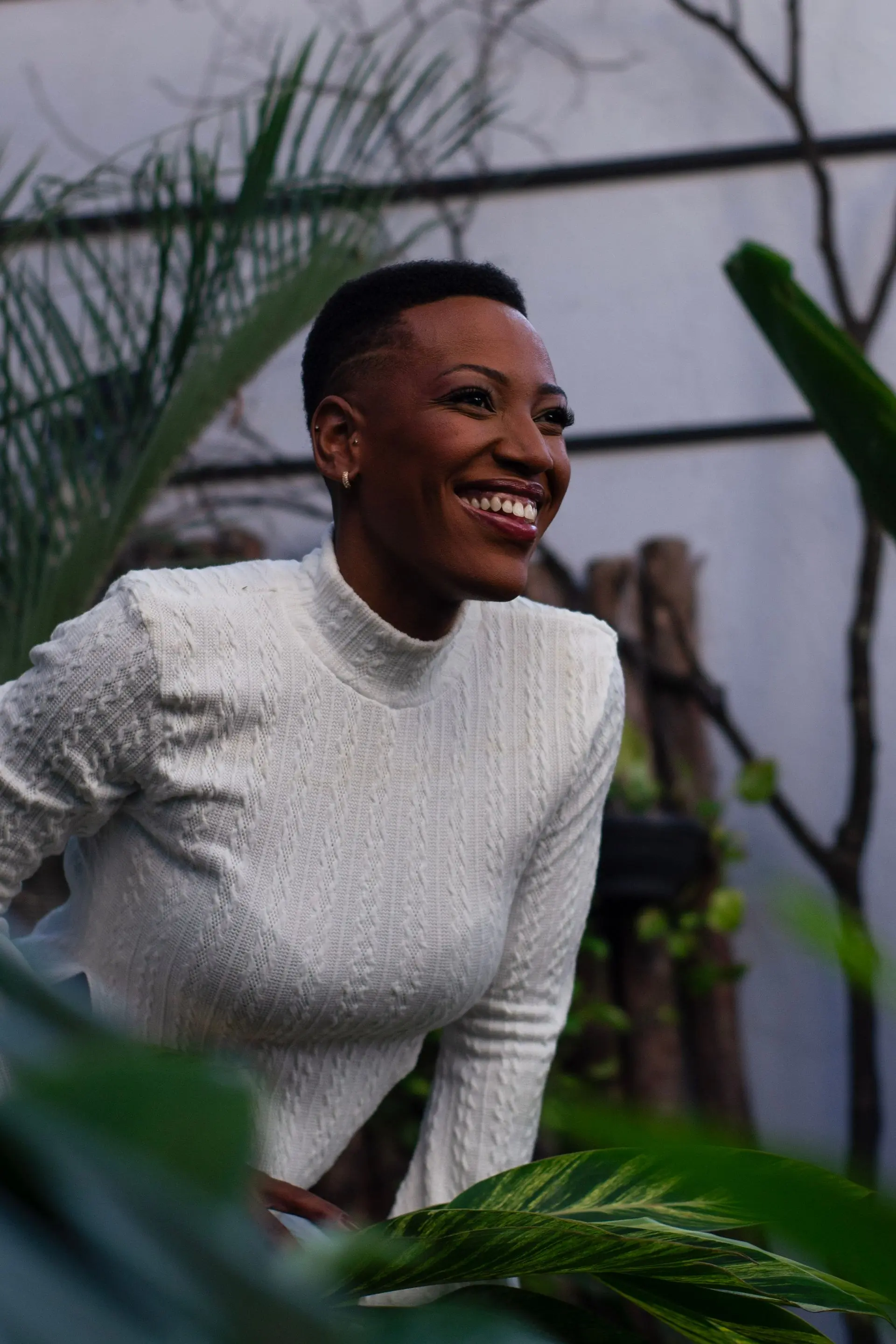 A woman in a white knit laughing among dark leaves