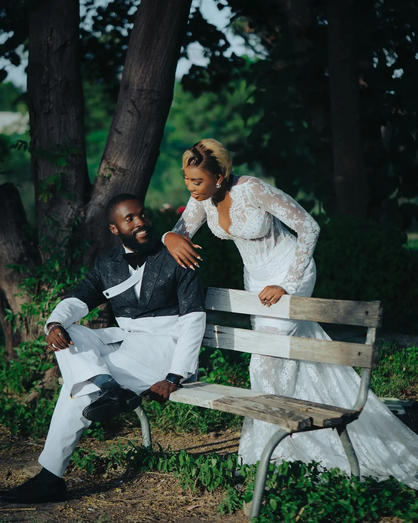 A couple in wedding clothes resting on a bench under a broad shady tree