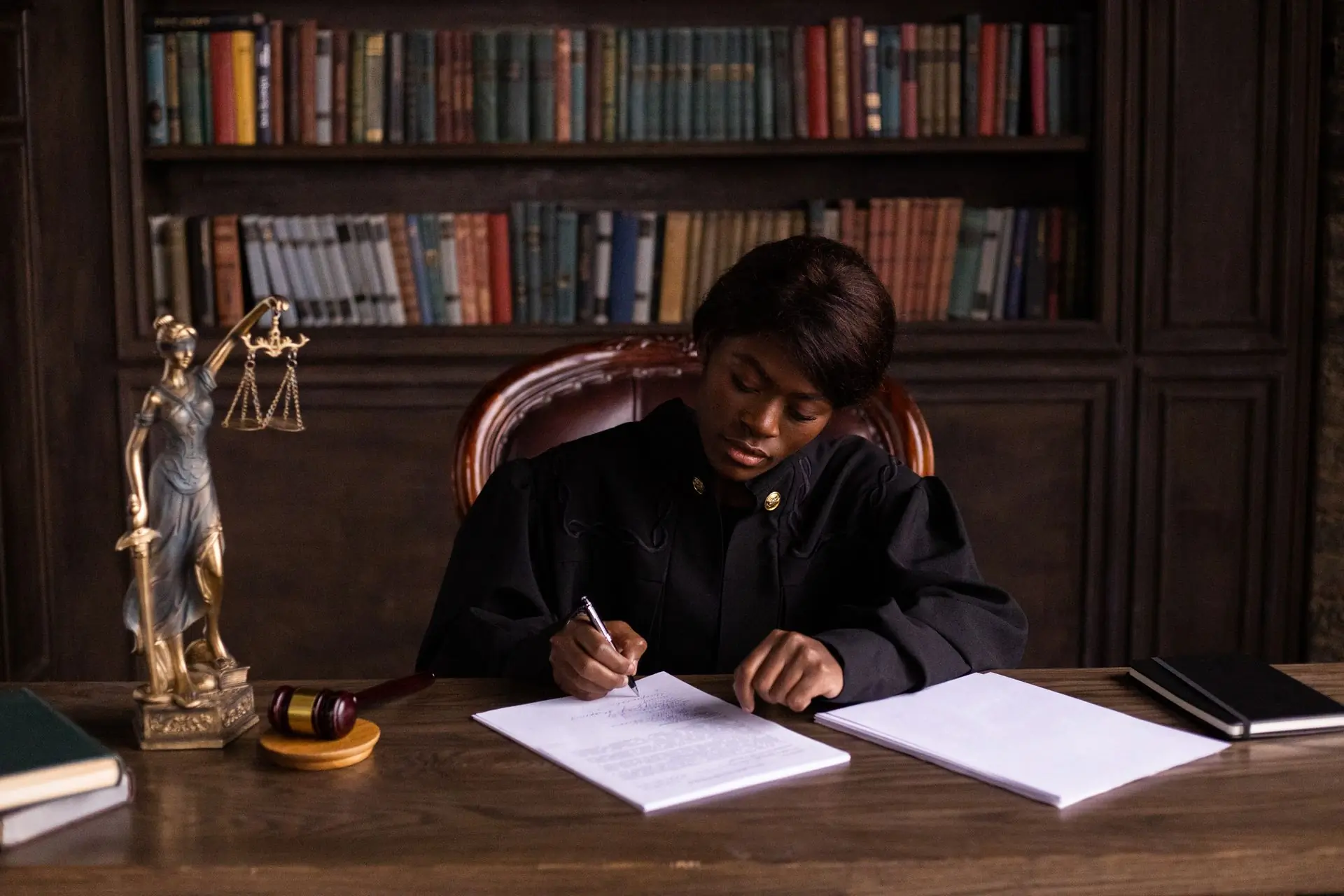 A judge writing at a desk in a dark, book-lined chamber