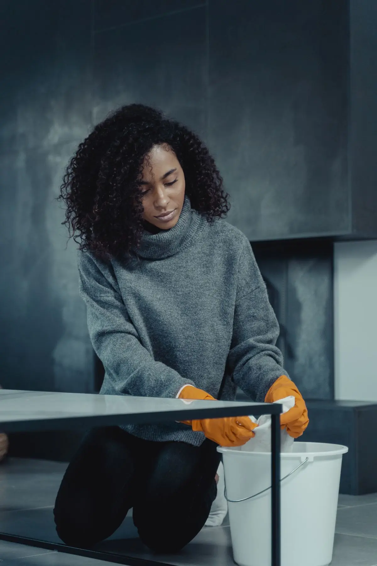 A cleaner working through a room, gloves on, bucket beside her