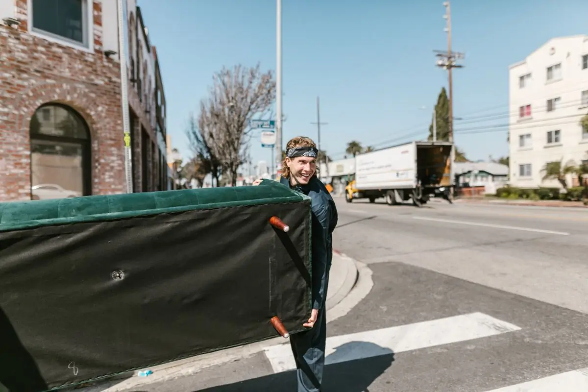 Two movers carrying a wrapped sofa across the street to a waiting van