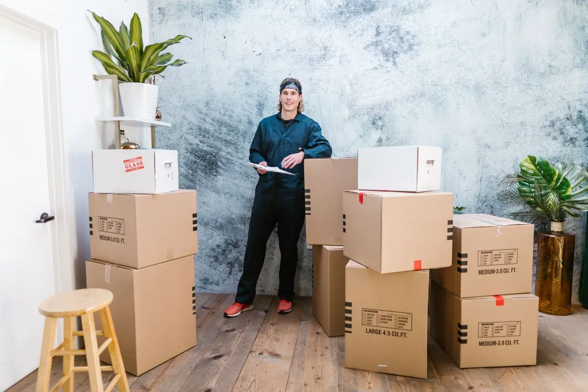 A packer in company overalls with a clipboard beside a stack of cartons