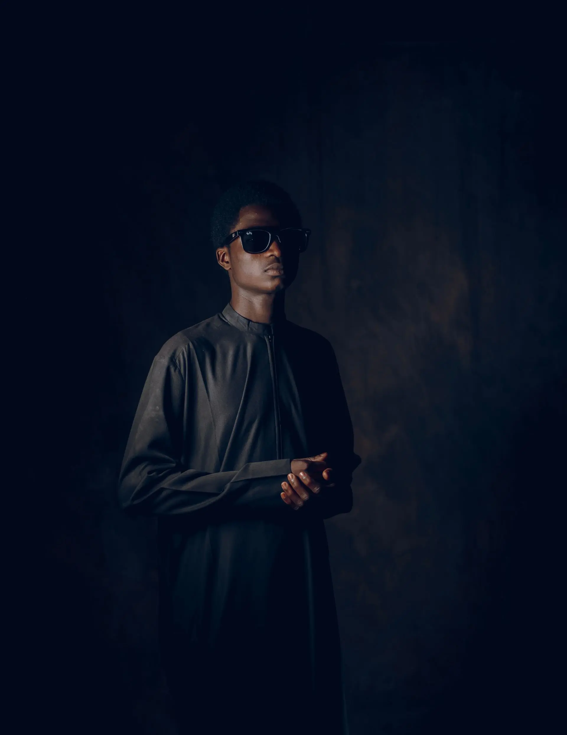 A man in a dark shirt and sunglasses photographed against a dark studio ground