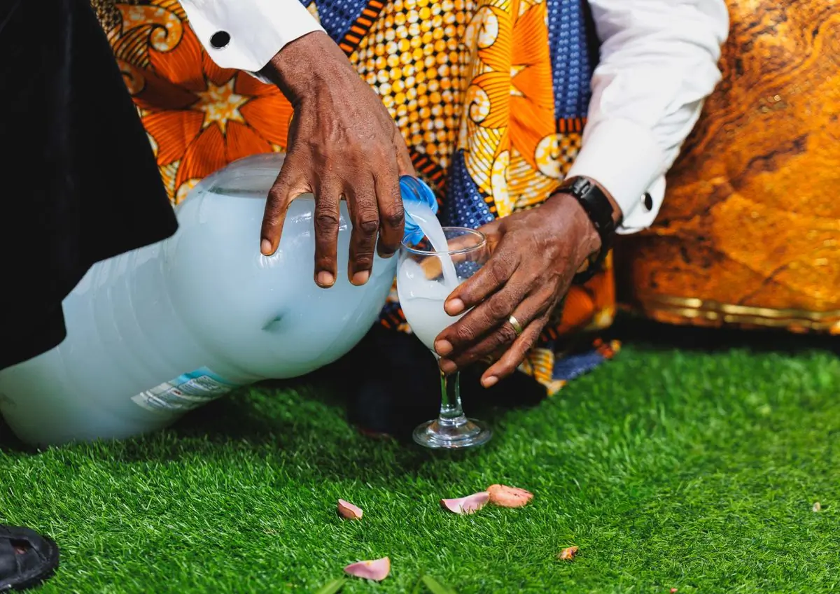 Hands pouring a libation into a glass over grass at an outdoor ceremony
