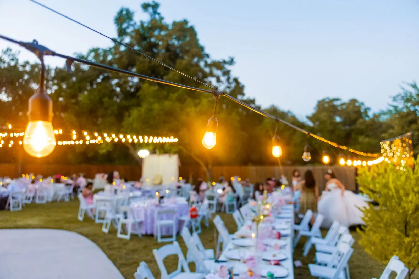 An outdoor reception at dusk lit by strings of bulbs above white chairs