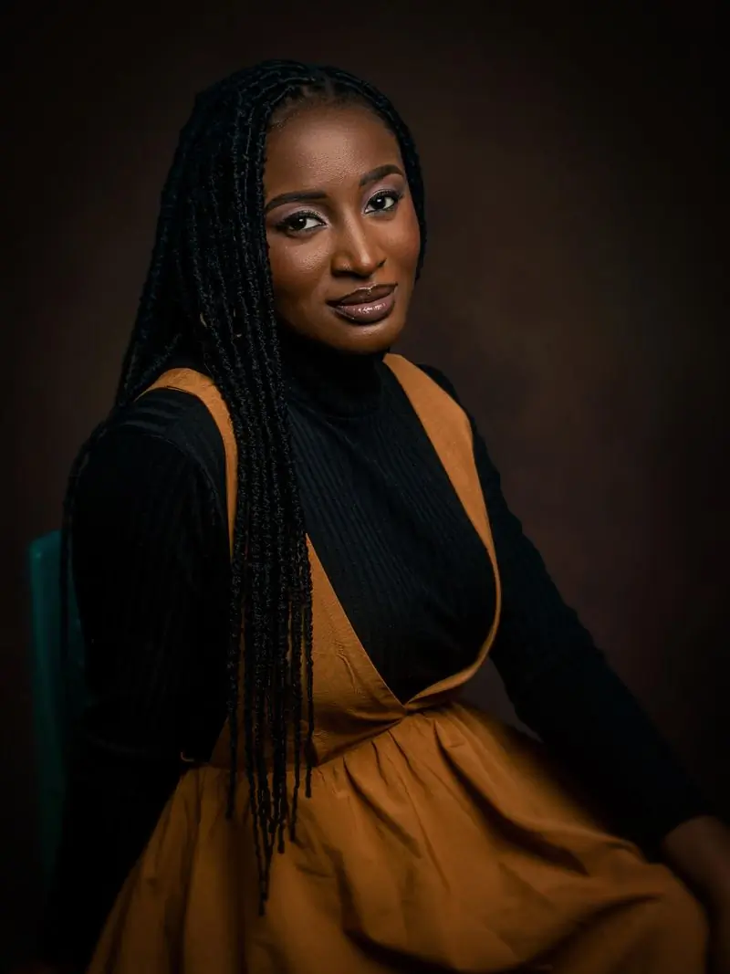 A woman with long braids in a mustard pinafore against a dark studio backdrop