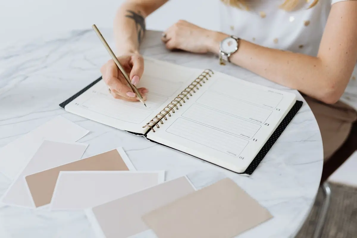 A planner's hands writing a running order in a notebook beside fabric swatches