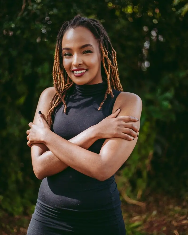A stylist with locs standing in front of a green hedge