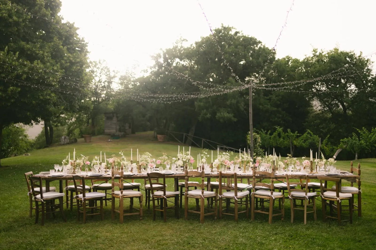 Long tables laid on grass under strung lights among mature trees at dusk
