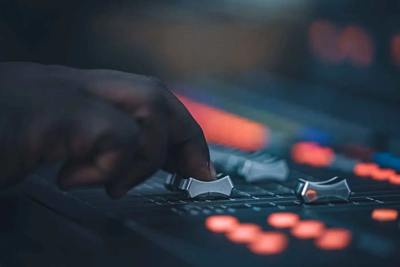 A hand adjusting faders on a lit mixing console in a dark room