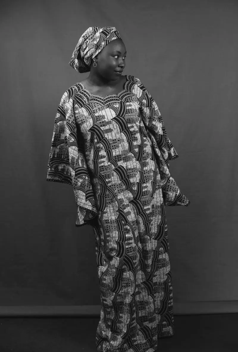 A woman in a patterned wax-print dress and headwrap, photographed in black and white