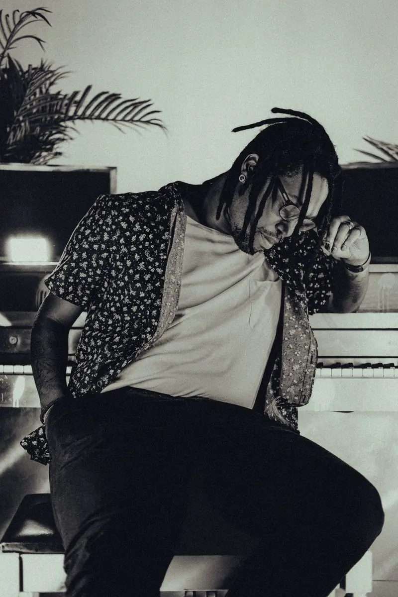 A man with locs leaning against an upright piano in a dim room