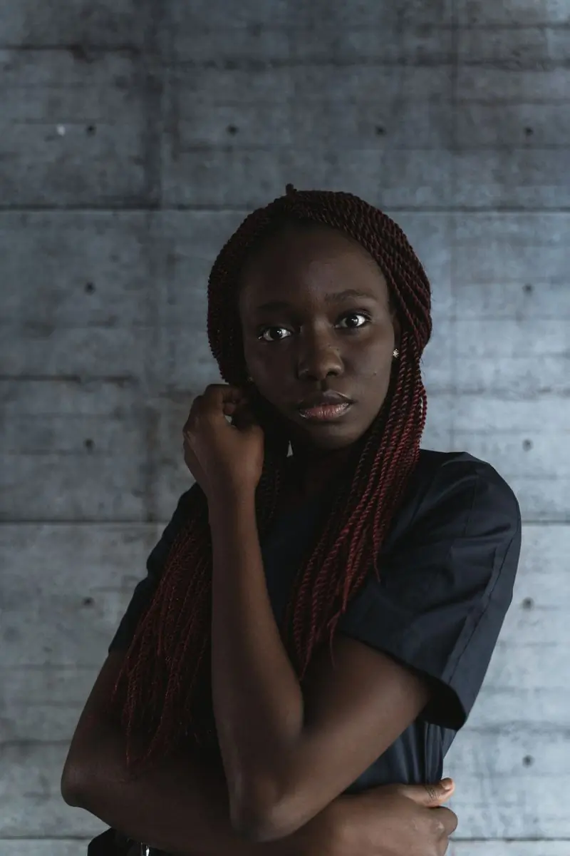 A woman with long braids standing against a concrete wall, looking straight ahead
