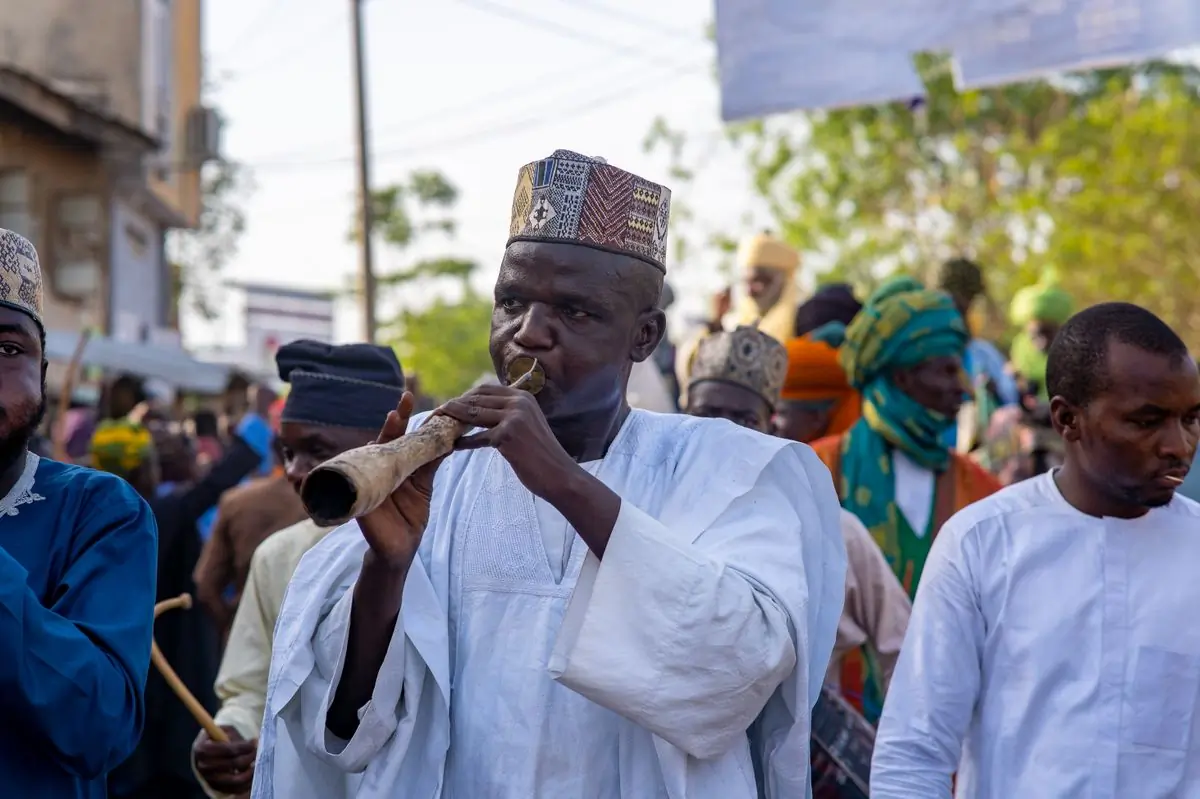 A man in white robes playing a long horn in a crowded procession