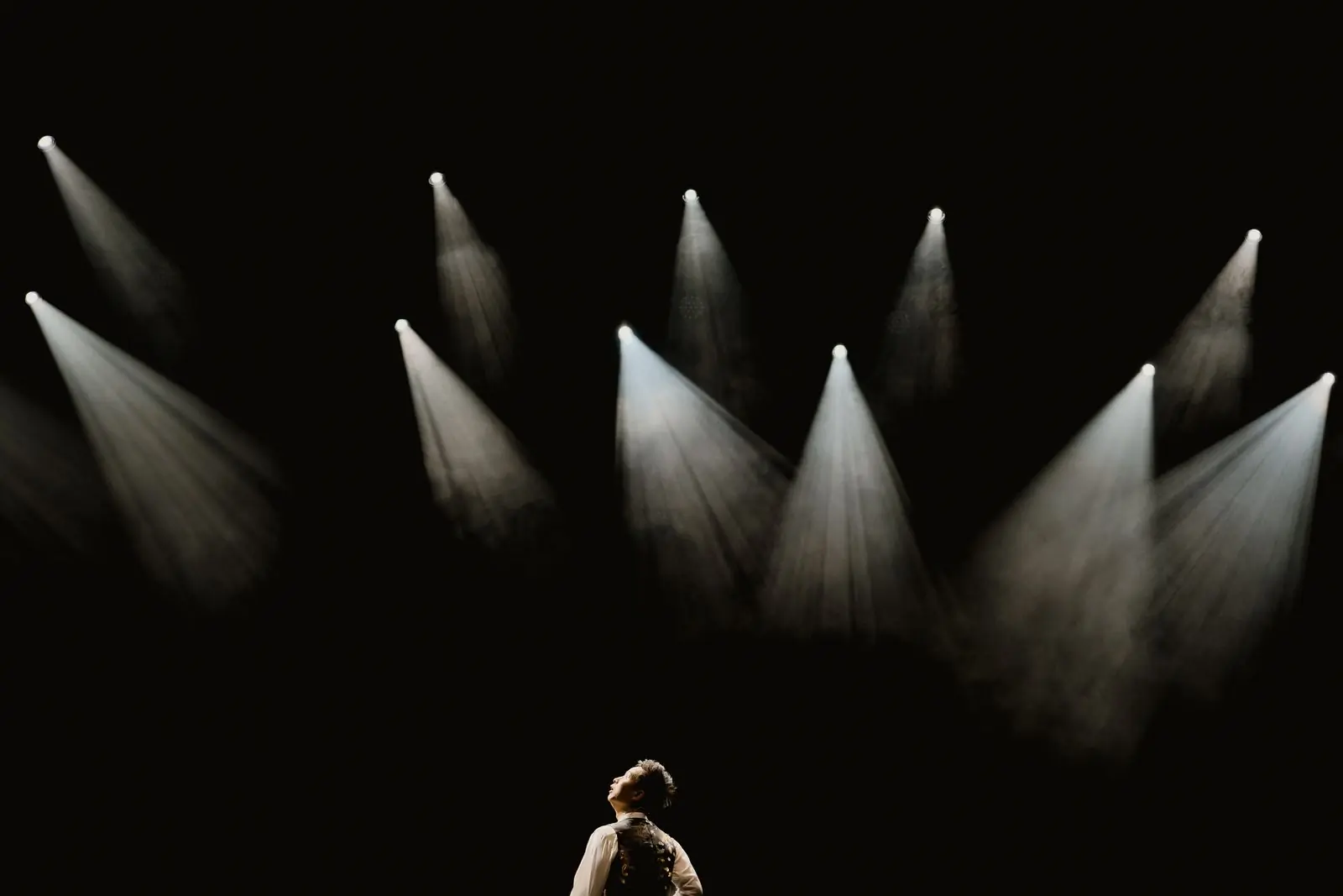 A performer alone on a dark stage beneath a bank of white spotlights