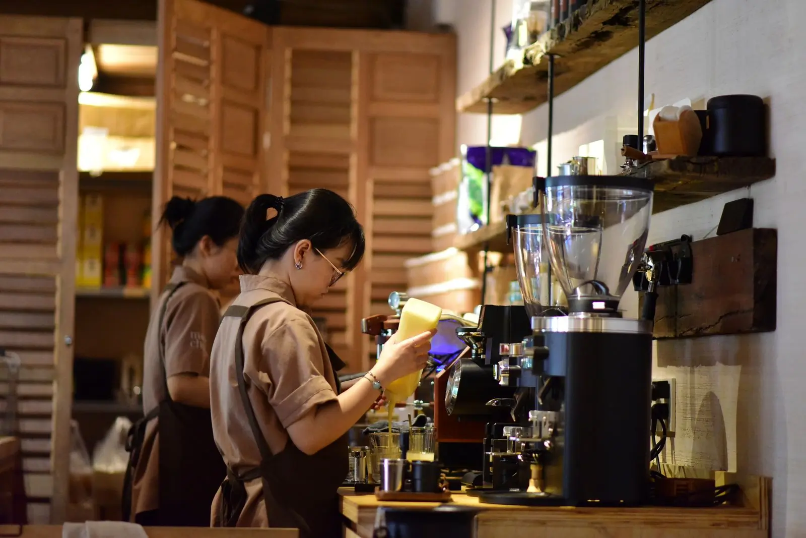 Two of the shop's baristas working side by side behind the counter, pouring and grinding under the lamps