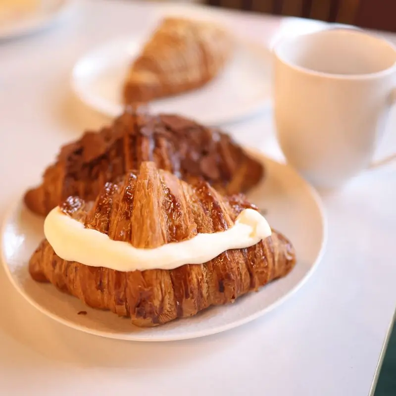 Two croissants on plates beside a cup of coffee on a bright table