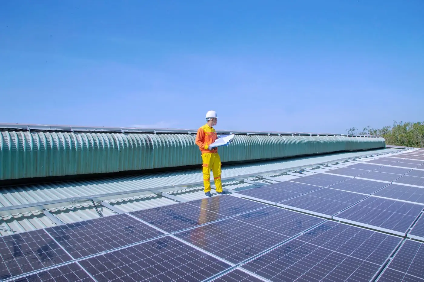 A long industrial roof covered in solar panels with a single technician walking the ridge