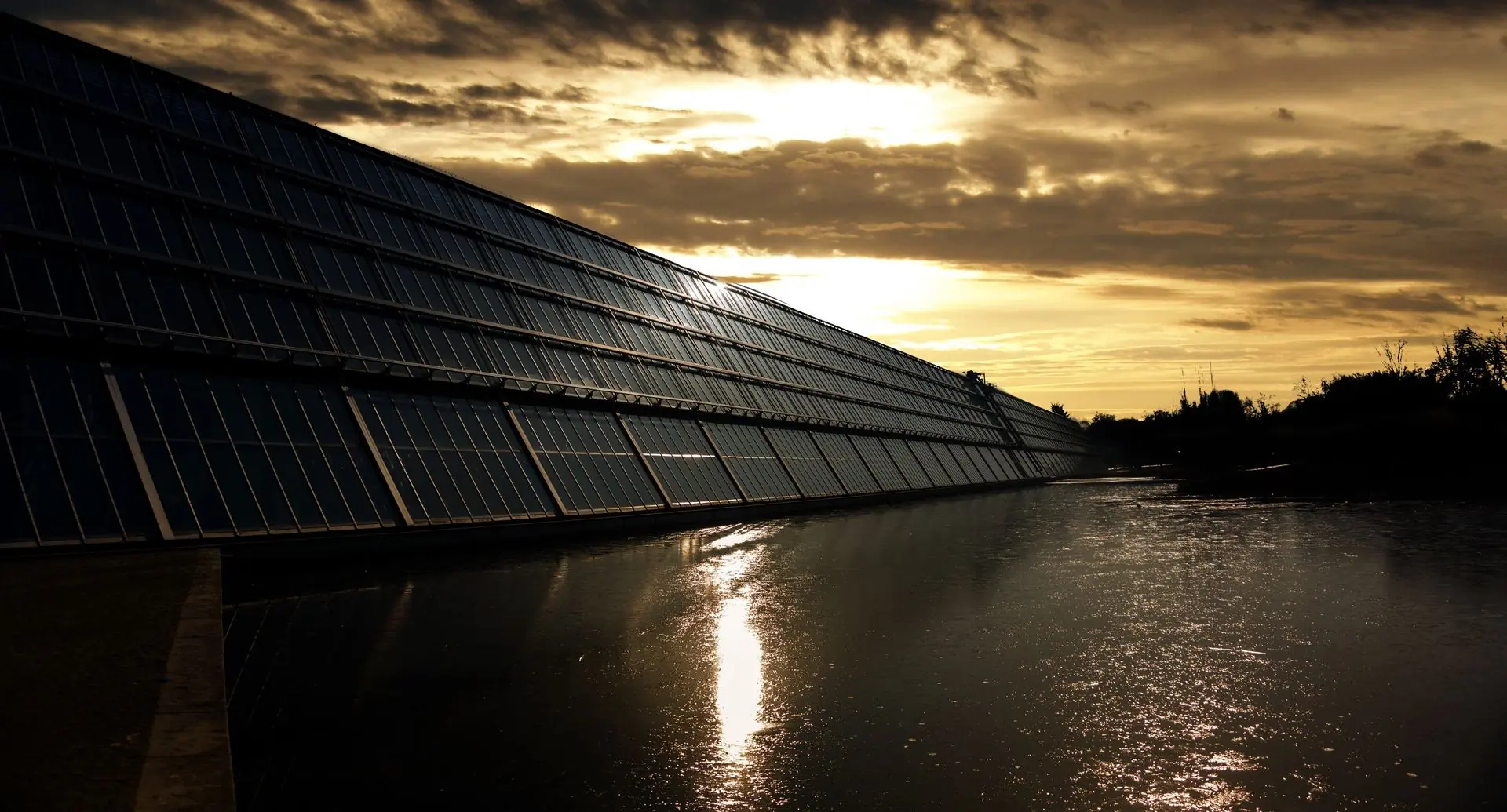 A long run of solar panels silhouetted against a low sun, reflected in still water below
