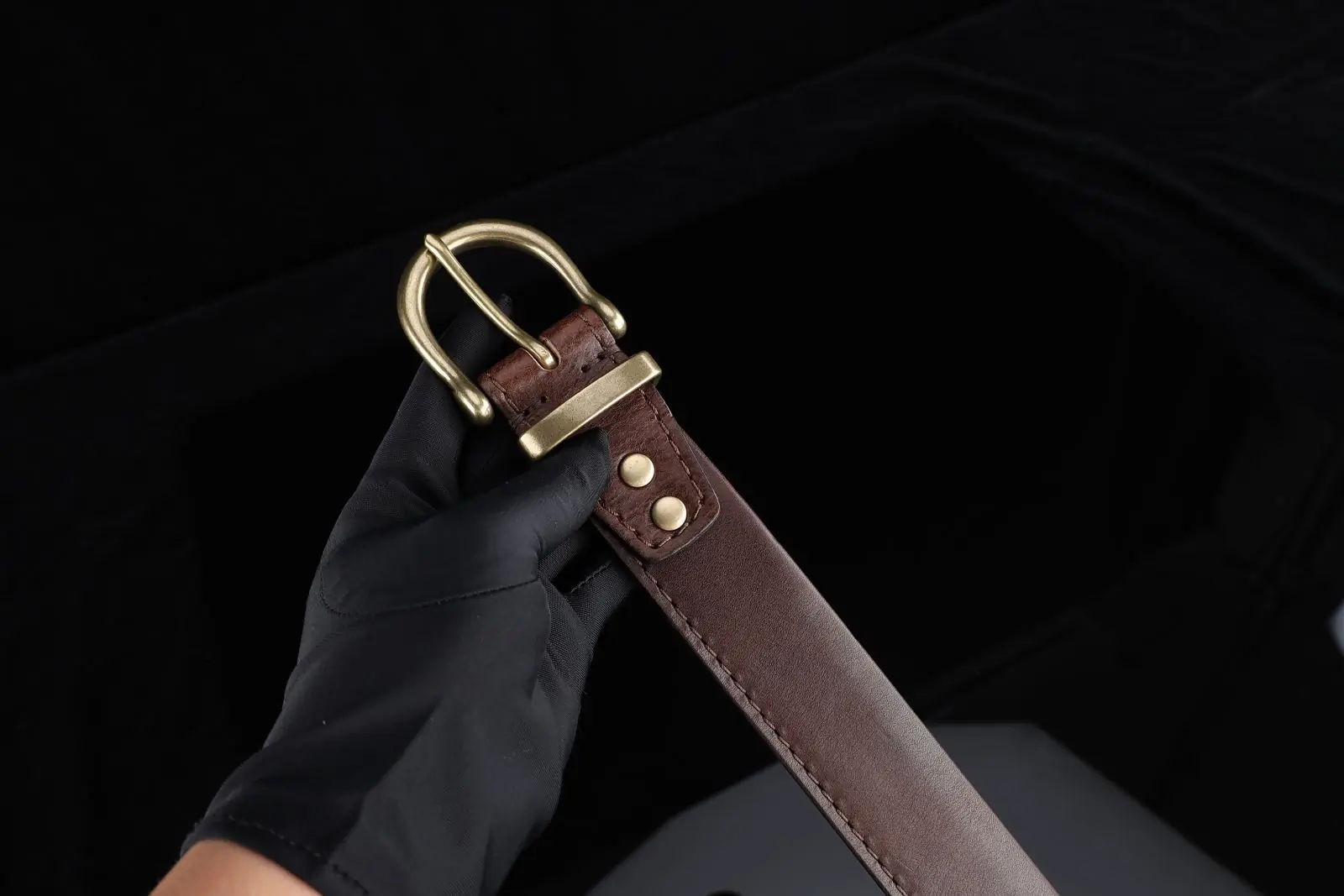 A gloved hand holding a brown leather belt with a brass buckle against black