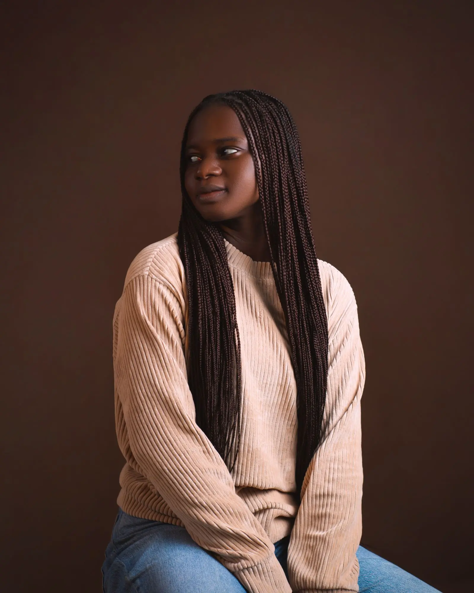 A woman with long braids in a blush ribbed knit, turned away against a deep brown ground