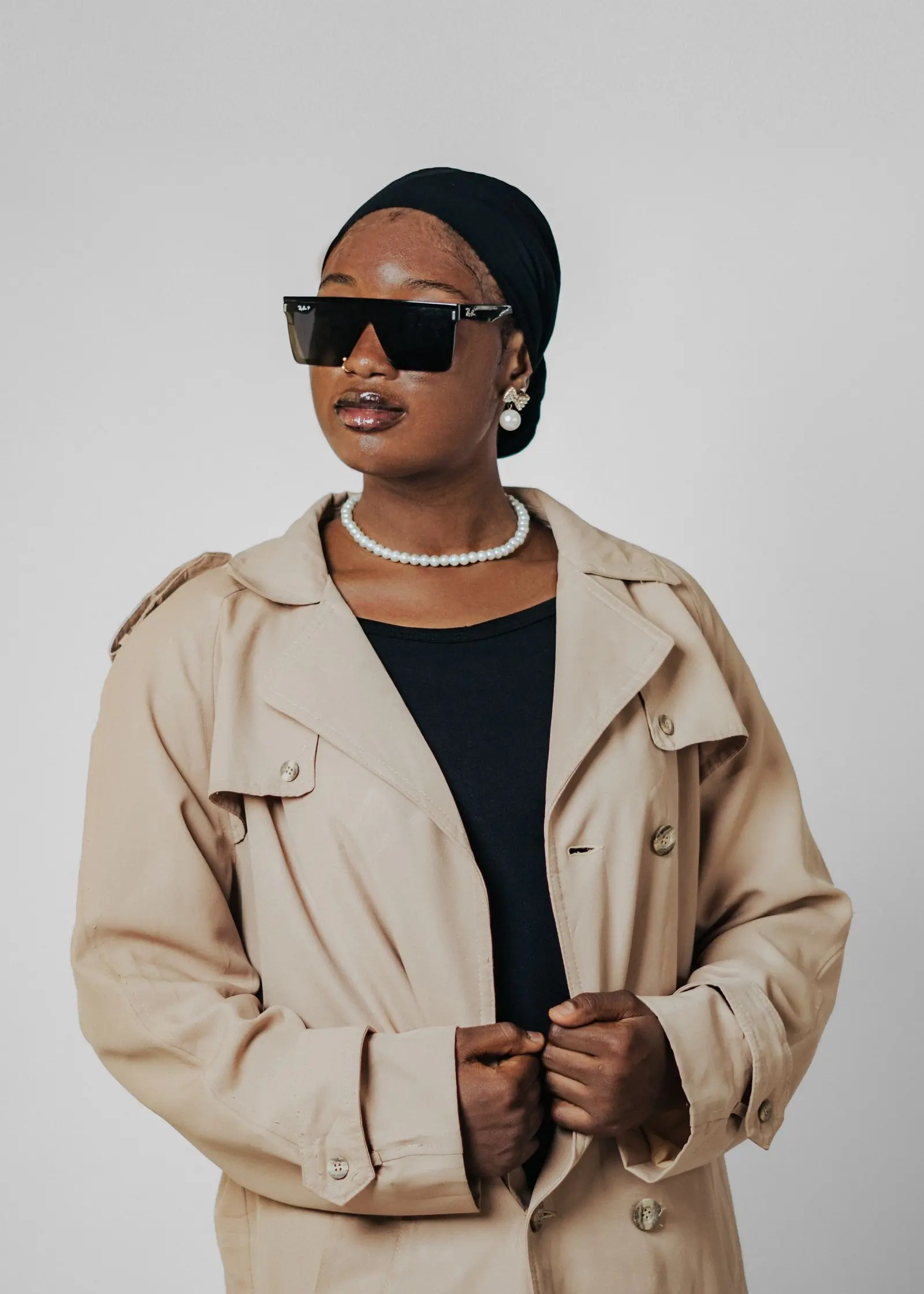 A woman in a stone trench coat and pearls, cropped at the chin against a pale wall