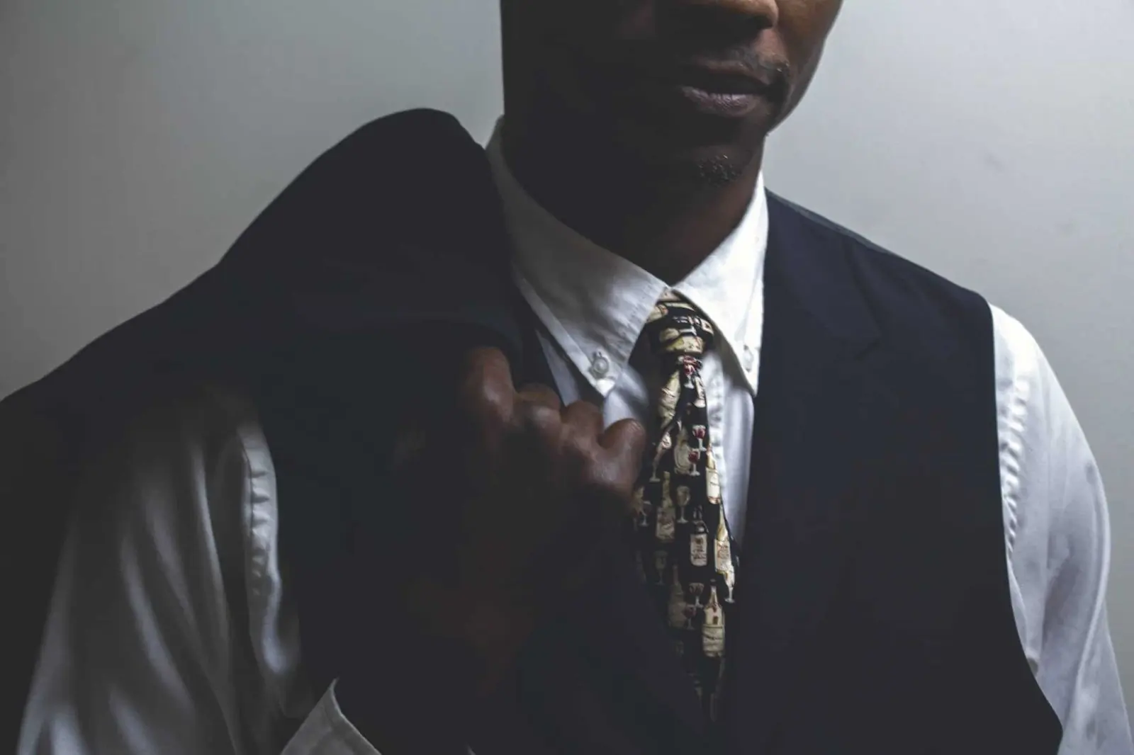 A man in a dark waistcoat, white shirt and patterned tie lit from one side in deep shadow