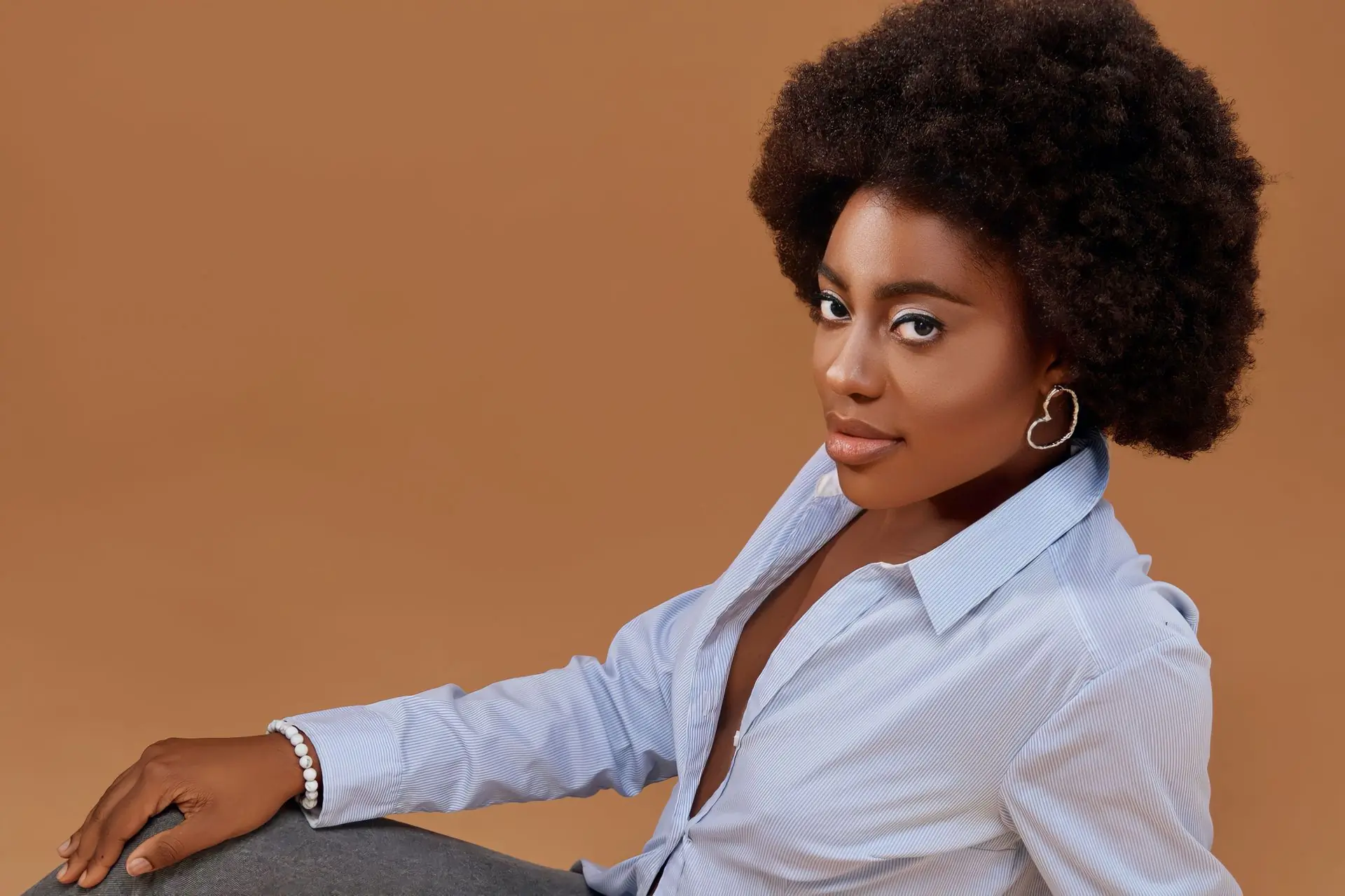 A woman with a full afro leans on a plinth in a pale striped shirt, lit against an empty camel studio wall