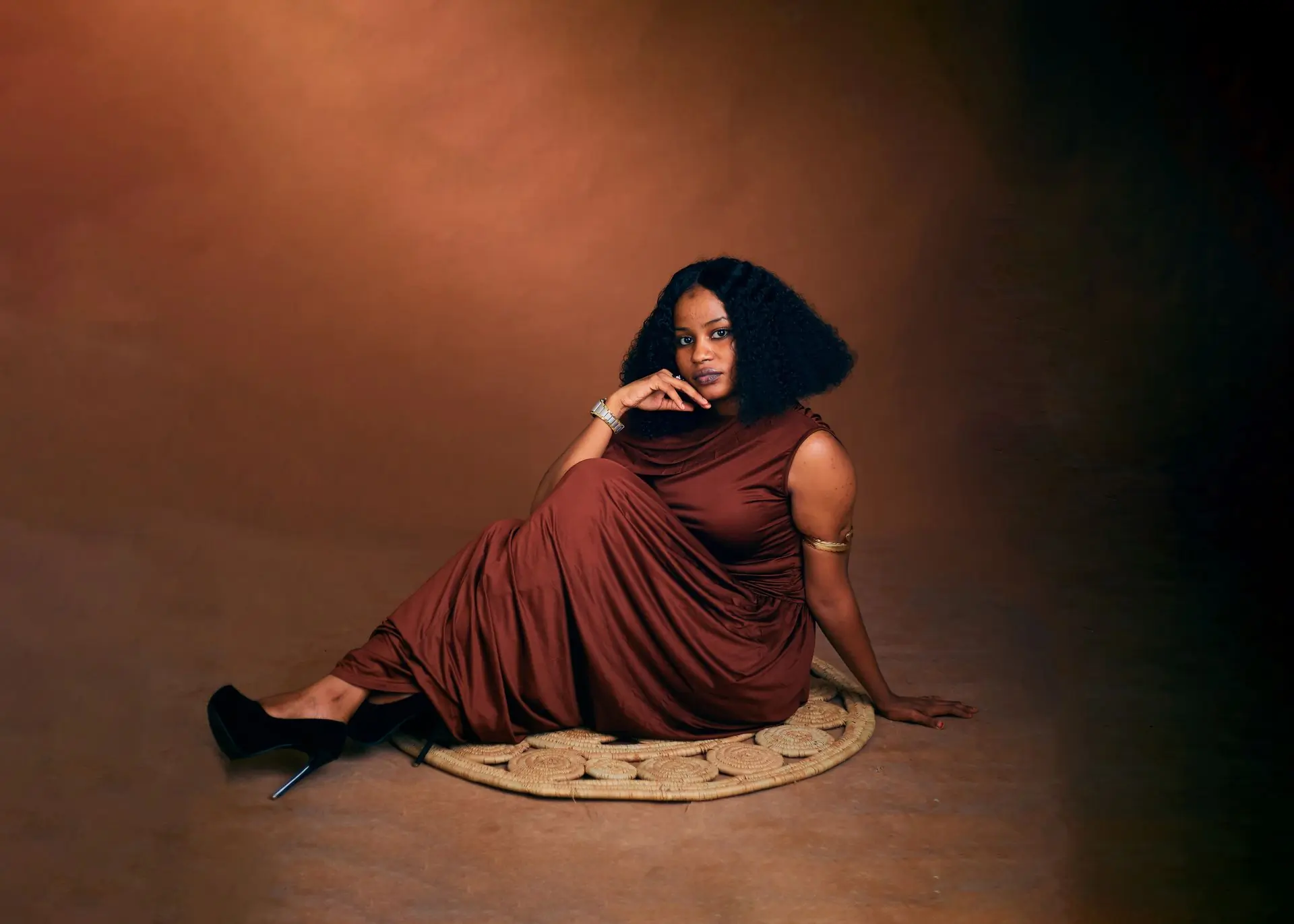 A woman in a draped rust-coloured dress reclines on a raffia mat against a mottled brown studio backdrop