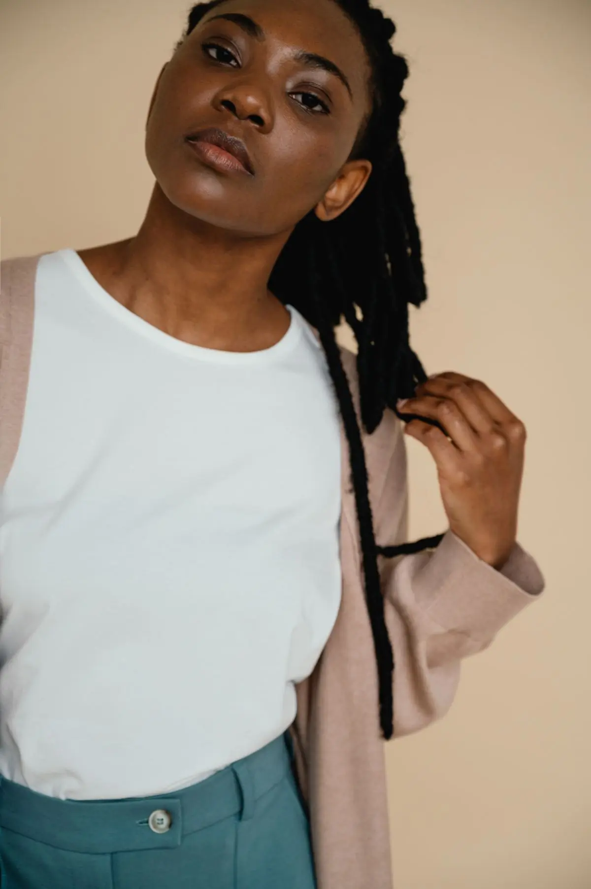 A woman with locs draws her hair aside over a white tank and blush jacket, cropped at the chin