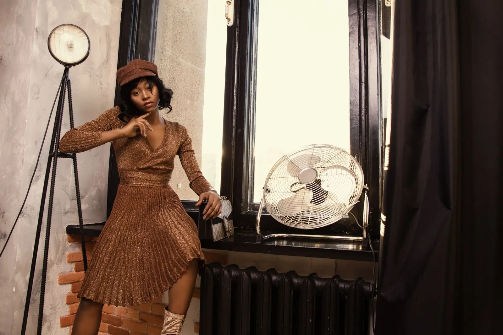 A woman in a bronze knit dress and cap leans on a window ledge in a warm, sunlit room
