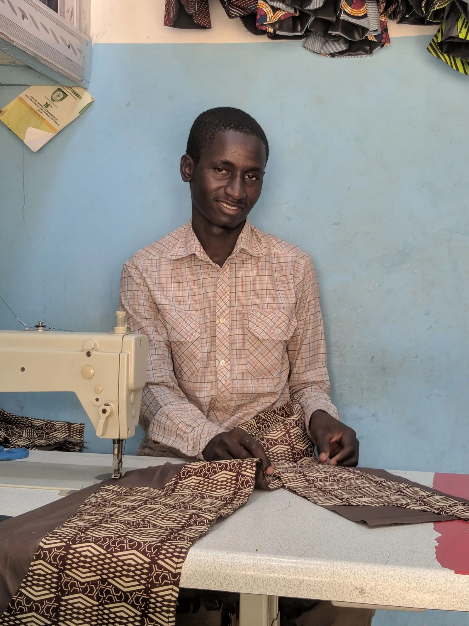 A tailor sits at his machine with cloth in his hands against a washed blue wall