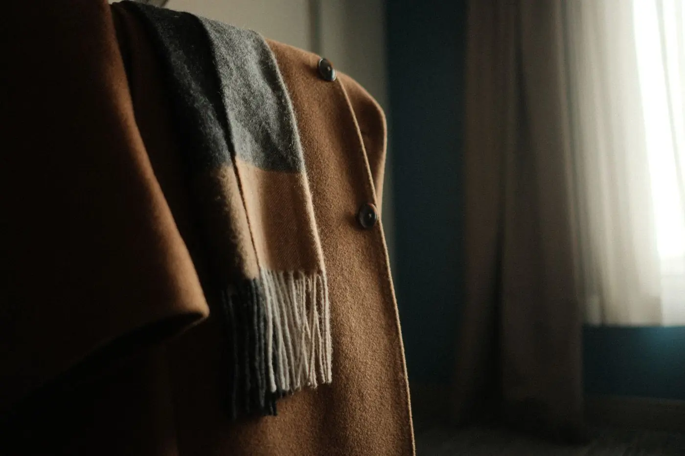 A camel wool coat with a fringed check scarf laid over the shoulder in low indoor light