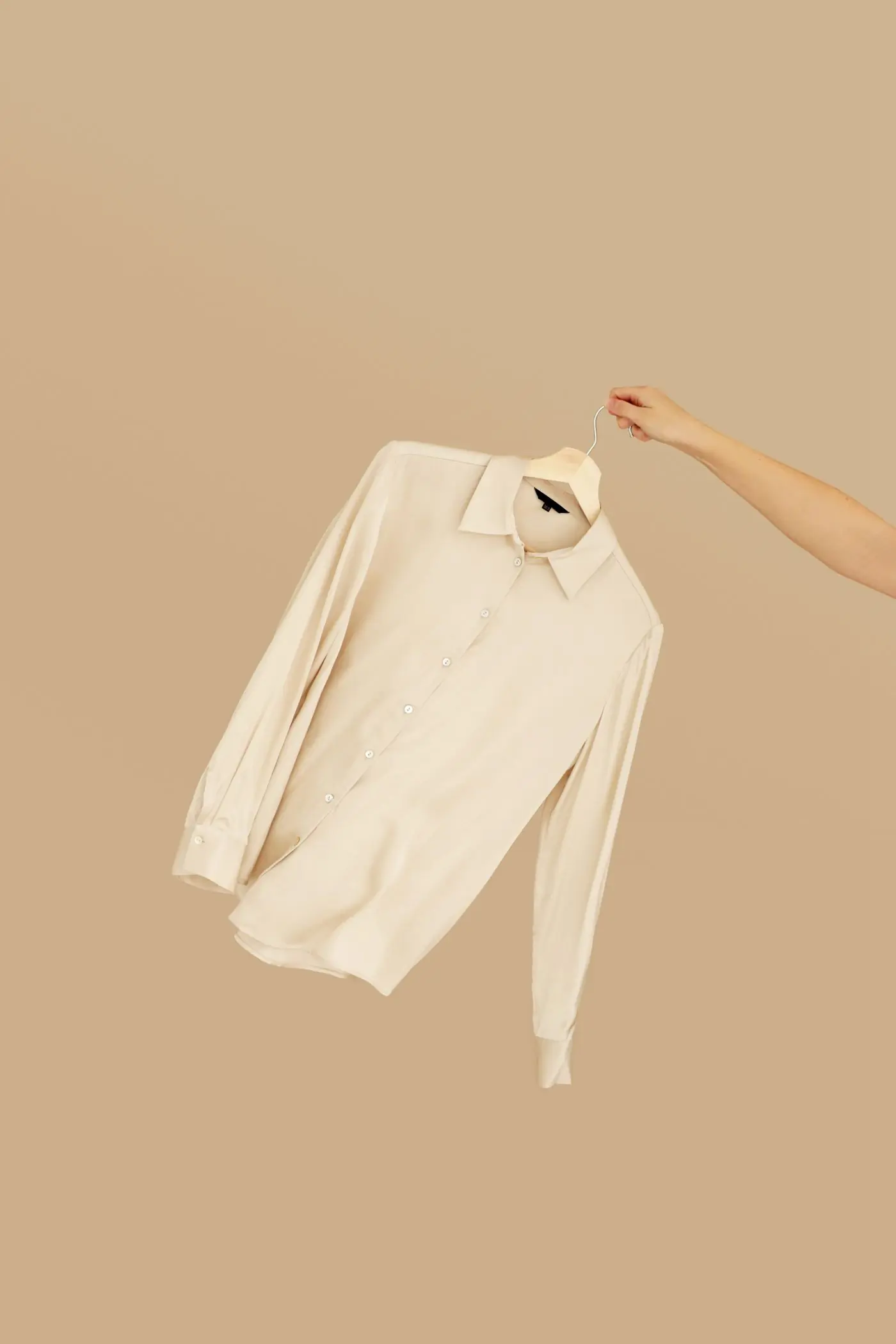 A cream silk shirt held up on a wire hanger against a camel backdrop
