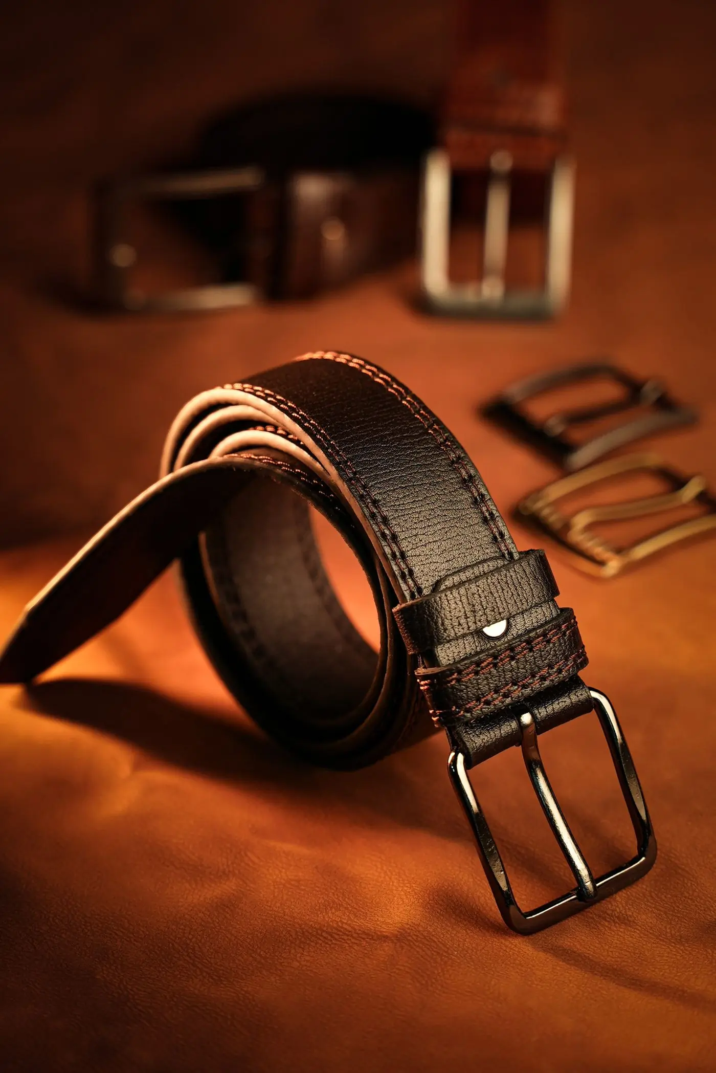A coiled dark leather belt with spare brass buckles laid on warm tan hide