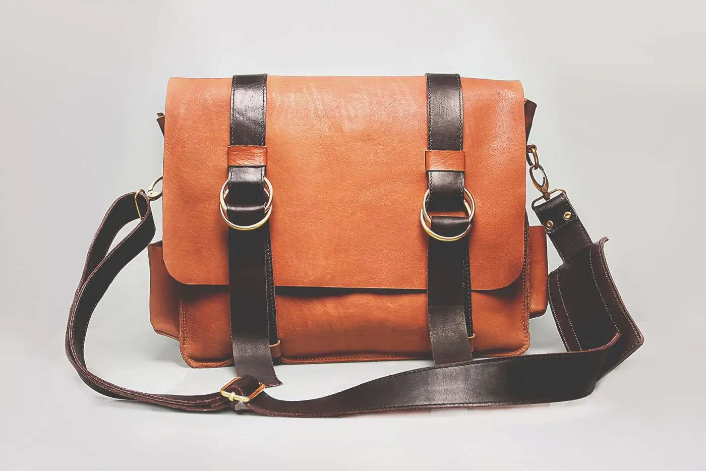 A tan leather satchel with dark buckled straps and a long shoulder strap, on white