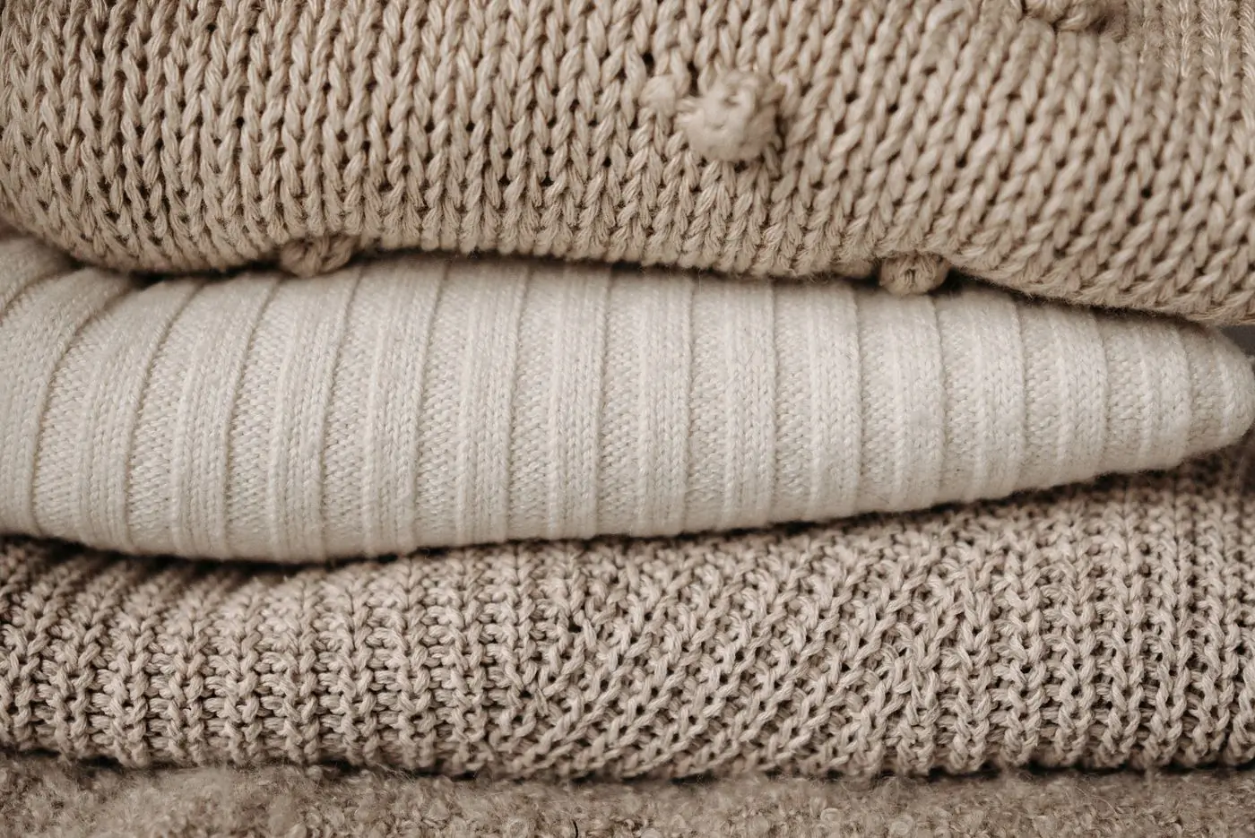 Three cream and oatmeal knitted sweaters folded in a stack, close up on the stitch