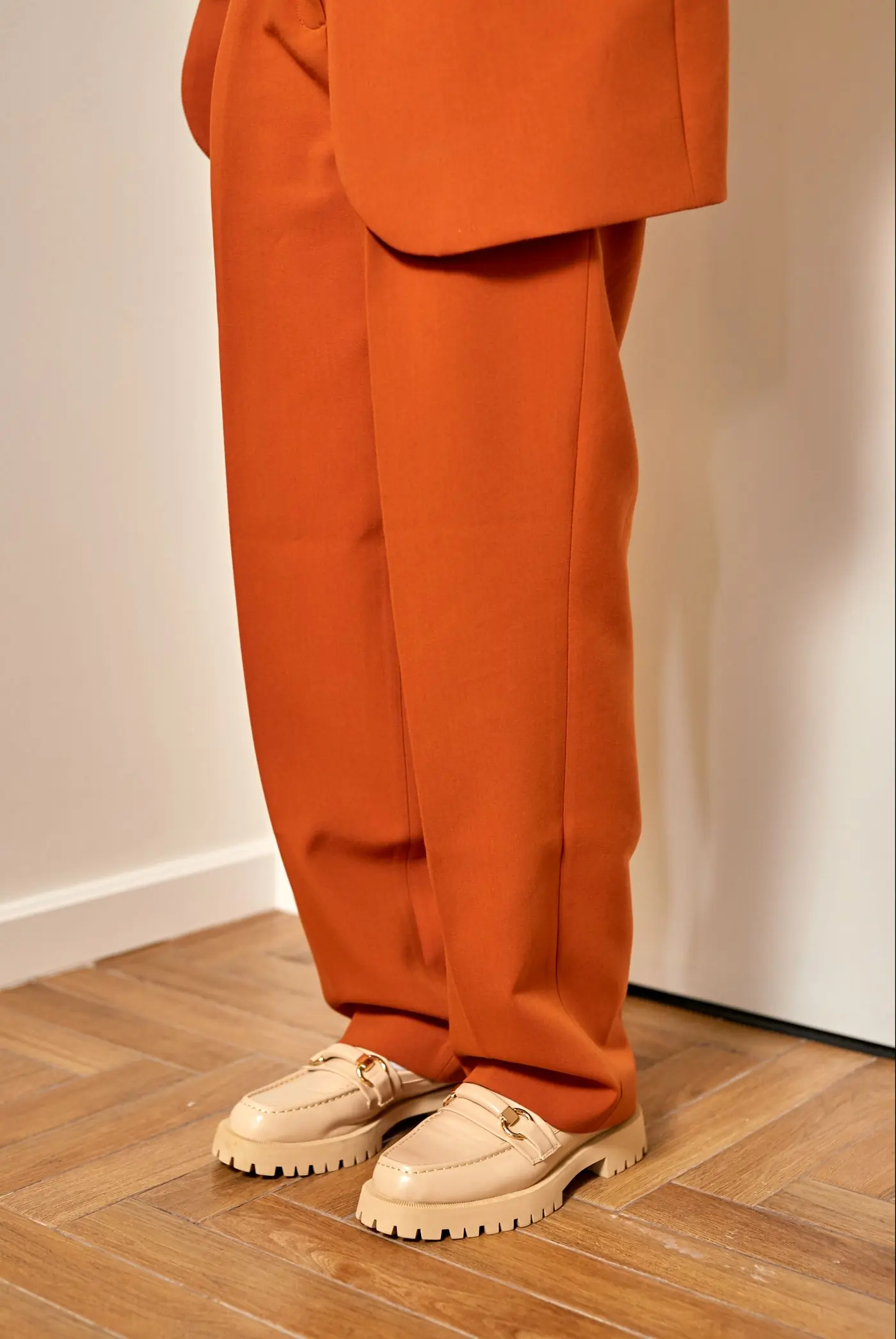 Pressed rust-coloured tailored trousers photographed against a white wall