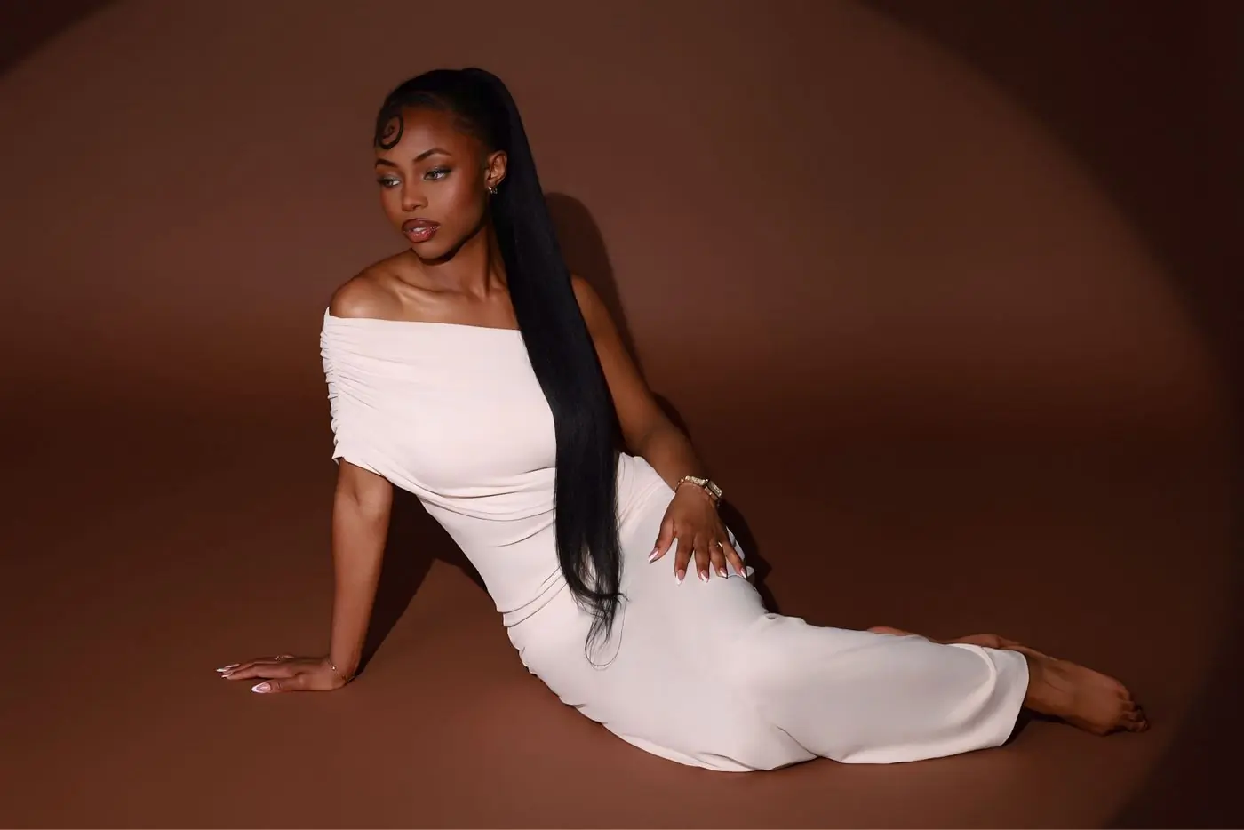 A woman in a long ivory off-shoulder dress seated on a warm brown studio floor