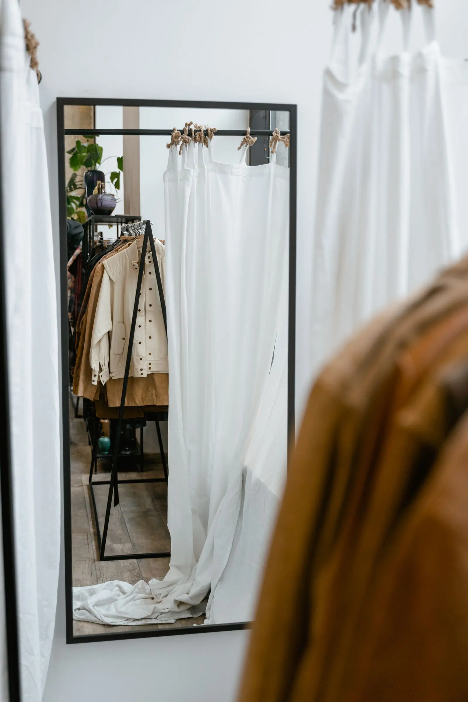 A mirror beside a linen curtain reflecting a rail of cream and tan garments