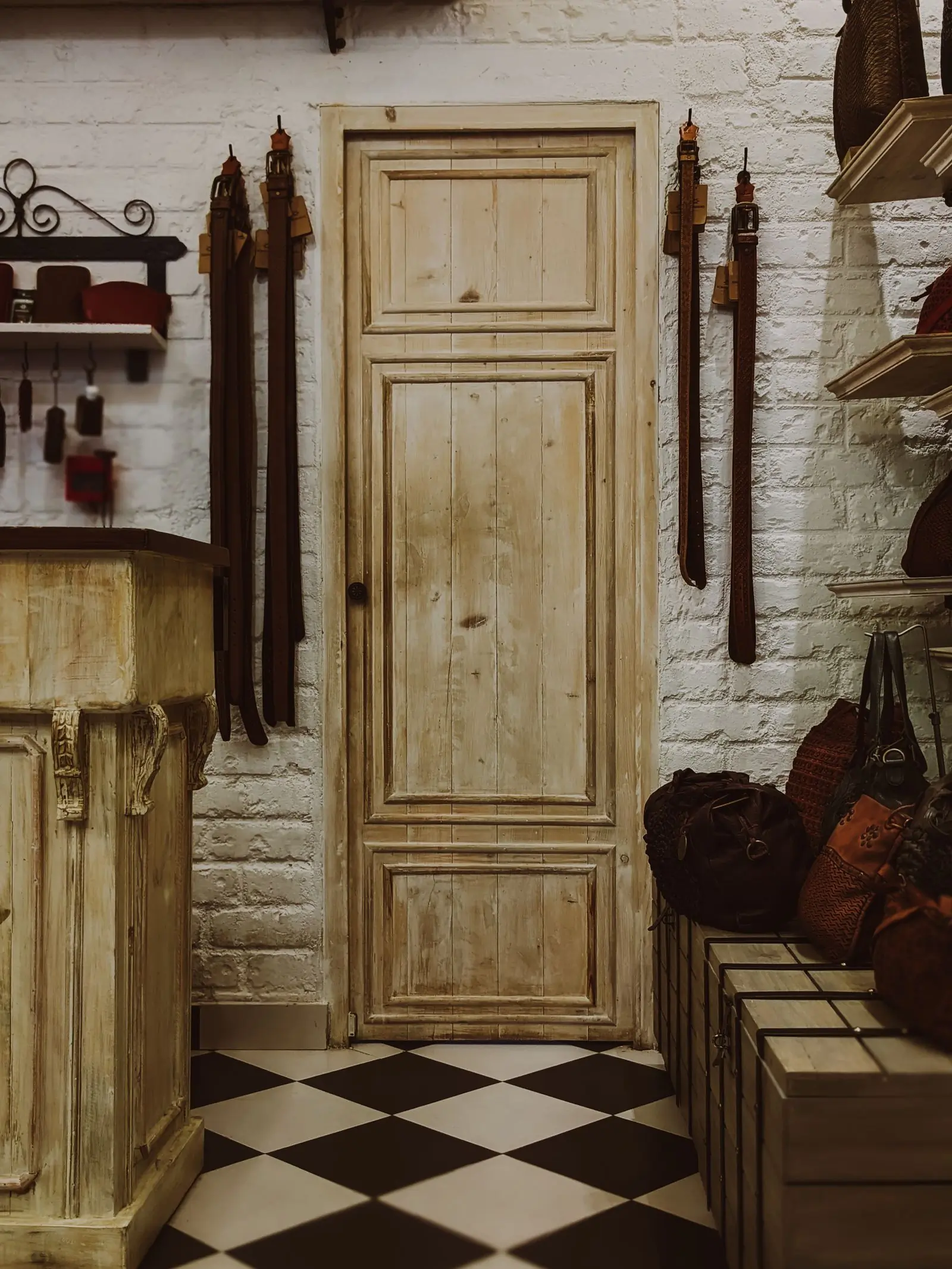Leather belts hung on a whitewashed brick wall beside a stripped wooden door and a pile of bags