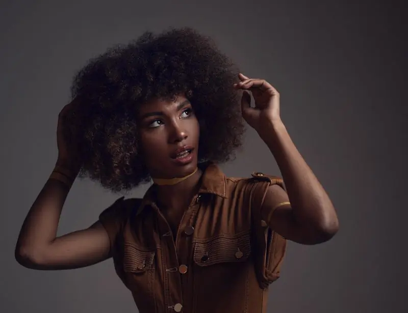 A woman with a full afro in a tobacco utility shirt against a grey-brown studio wall